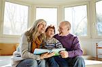 Grandparents on window seat reading book with grandson, smiling