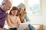 Grandparents on window seat using digital tablet with granddaughter looking down smiling