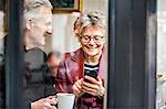 Senior couple sitting at cafe window seat drinking coffee and texting on smartphone