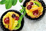 Cakes with slices of fresh fruit orange, kiwi and grapes with jelly and cream on a black saucers on a table cloth.