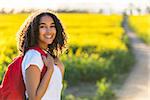 Outdoor portrait in golden evening sunshine of beautiful happy mixed race African American girl teenager female young woman smiling laughing with perfect teeth hiking with red rucksack in yellow field