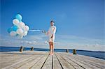 Portrait of young woman, standing on wooden pier, holding bunch of balloons