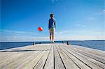 Young boy jumping on wooden pier, holding red helium balloon, rear view