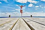 Young woman dancing on wooden pier, mid air, rear view