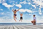 Young woman sitting on post on wooden pier, watching friends jump into sea, rear view