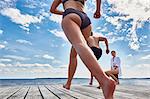 Young woman sitting on post on wooden pier, watching friends running towards edge, low angle view