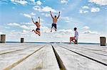 Young woman sitting on post on wooden pier, watching friends as they jump into sea