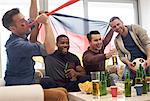 Group of men watching sporting event on television holding German flag and football, celebrating