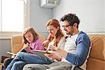 Mid adult parents and daughter on sofa concentrating on reading book and digital tablet