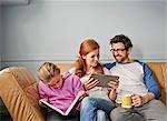 Mid adult parents and daughter on sofa on reading book and digital tablet