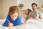 Mid adult couple having lie in whilst daughter draws on bed