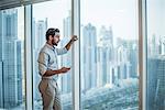 Businessman with smartphone staring through window with skyscraper  view, Dubai, United Arab Emirates