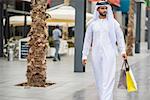 Man wearing traditional middle eastern clothing walking along street carrying shopping bags, Dubai, United Arab Emirates