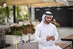 Male shopper  wearing traditional middle eastern clothing sitting on bench reading smartphone text, Dubai, United Arab Emirates