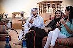 Local couple wearing traditional clothes smoking shisha on sofa with female tourist, Dubai, United Arab Emirates