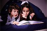 Brother and sister under duvet reading book by torchlight