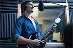 Male musician in recording studio, singing into microphone and playing guitar