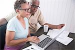 Senior couple sitting at table, using laptop, looking at paperwork