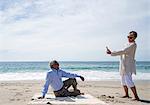 Senior couple on beach, woman taking photograph of man using smartphone