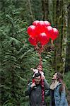 Parents strolling with toddler daughter on shoulders and bunch of red balloons in forest