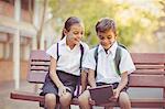 Happy school kids sitting on bench and using digital tablet