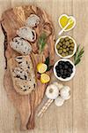 Olive bread on a wooden board with black and green olives and leaf sprigs, with lemon, oil and garlic bulbs over oak background.