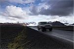 Horizontal photo of an off road car on a straight asphalt road coming from the mountains with clouds above and the Vatnajokull glacier in the background, Iceland