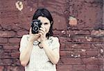 Girl holding retro camera and taking photo on vintage brick wall background. Selective focus on camera and hands of model. Toned photo with copy space. Vintage style photo.