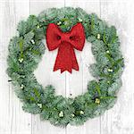 Christmas wreath with red bow decoration, mistletoe and snow covered blue spruce fir over distressed white wood background.