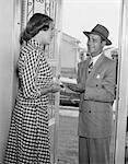 1950s HOUSEWIFE IN GINGHAM DRESS AT DOOR OF HOME TALKING TO SALESMAN IN SUIT & HAT HOLDING PENCIL & PAD