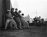 1950s TWO GROUPS OF FIVE MEN EACH PULLING ON TUG-OF-WAR ROPE ON BUILDING ROOFTOP TRYING TO PULL SOMETHING APART