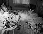 1950s SAD BLOND WOMAN LOOKING STOOD UP WEARING CHIFFON RUFFLED EVENING GOWN LYING ON BED WAITING BY TELEPHONE