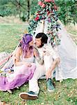 Sweden, Bride and groom sitting on grass by white tent at hippie wedding