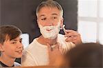 Son watching father shave face in bathroom mirror