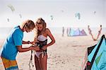 Man fastening kiteboarding safety harness onto woman on sunny beach