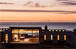 Illuminated luxury home overlooking ocean at sunset