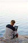 Sweden, Ostergotland, Teenage boy (16-17) sitting on rock and using digital tablet