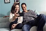 Sweden, Young couple sitting on sofa, looking at smart phone and laughing