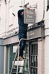 A man on a ladder fixing a painted name sign onto a bracket on a shopfront.