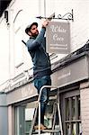 A man on a ladder fixing a painted name sign onto a bracket on a shopfront.