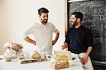 Two bakers standing at a table, using a laptop computer, freshly baked bread, a blackboard on the wall.