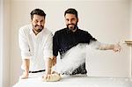 Two bakers standing at a table, kneading bread dough, dusting it with flour.