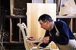 Man standing in a carpentry workshop, working on a wooden chair marking the armrest joint with a pencil.