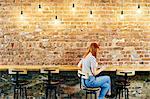 Female customer sitting at brick wall bench browsing digital tablet in coffee shop