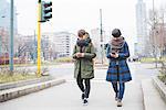 Two sisters walking along street, using smartphones