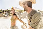 Mid adult couple wearing straw hat and feather headdress running on beach, Cape Town, South Africa