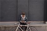 Young man sitting on wall, bike in front of him, using smartphone