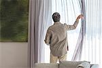 Rear view of a man holding curtain on window