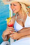 Stunningly beautiful young blond woman in straw cowboy hat and white bikini enjoying a cocktail by a turquoise blue sea