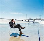 Businessman in deckchair on Los Angeles river using smartphone and laptop, California, USA